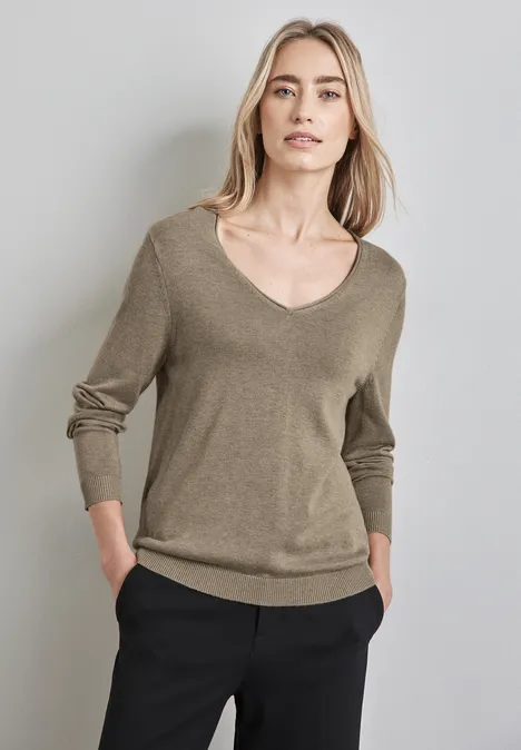 Frau mit hellbraunem Pullover und schwarzer Hose steht vor hellem Hintergrund.
